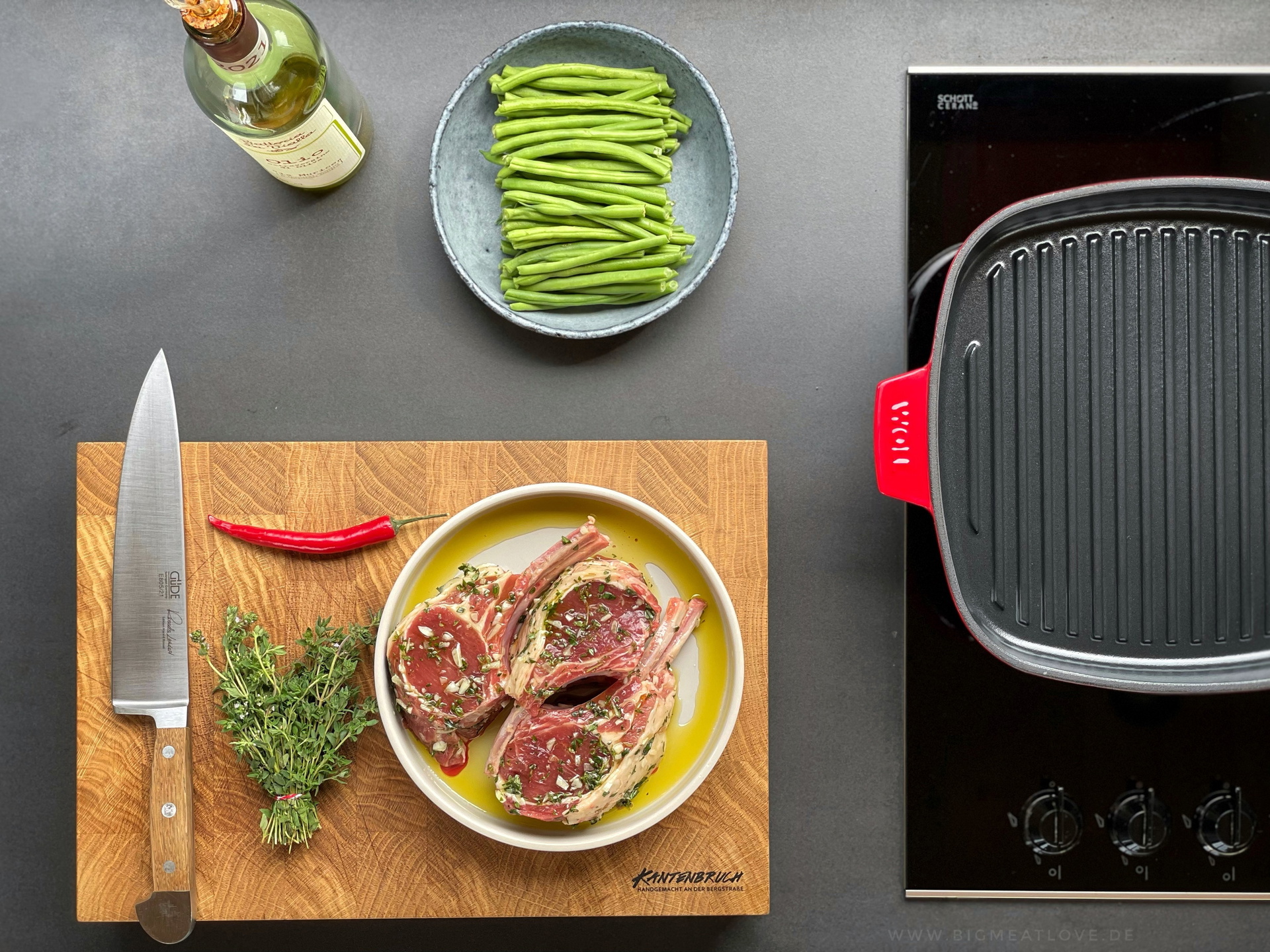 Auf dem Herd steht eine viereckige Grillpfanne der Serie Iron, auf dem Holzbrett daneben liegen ein Messer, Kräuter und ein Teller mit Lammkarree in eingelegter Marinade. Darüber steht eine Schüssel mit Bohnen.