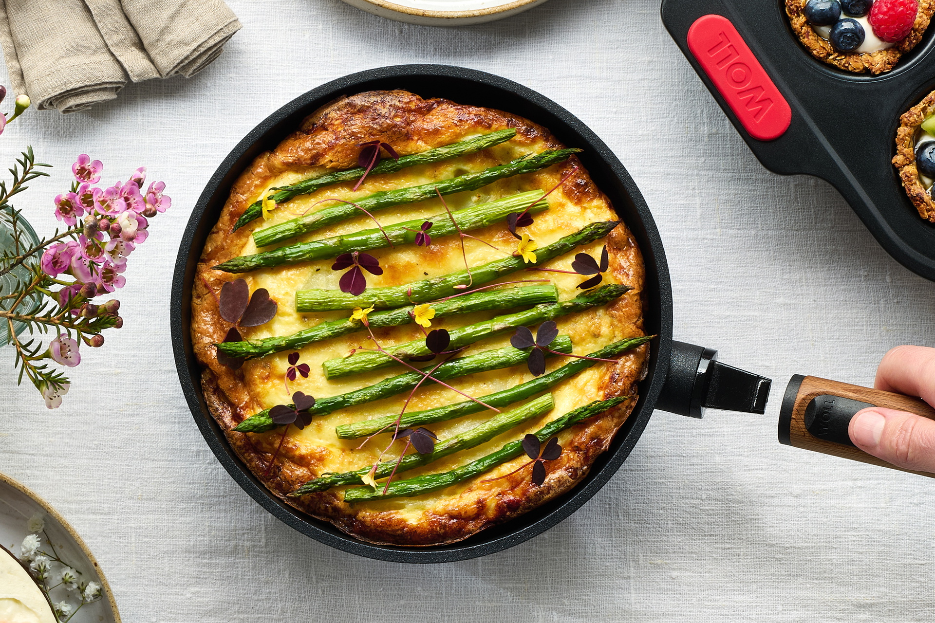 In einer runden Pfanne der Serie Eco Logic QXR befindet sich ein frisches Kartoffel-Spargel-Omelet. Der Holzstiel der Pfanne wurde abgenommen, damit sie auf den Tisch gestellt werden kann. Daneben steht eine Muffinform mit Granola-Cups.