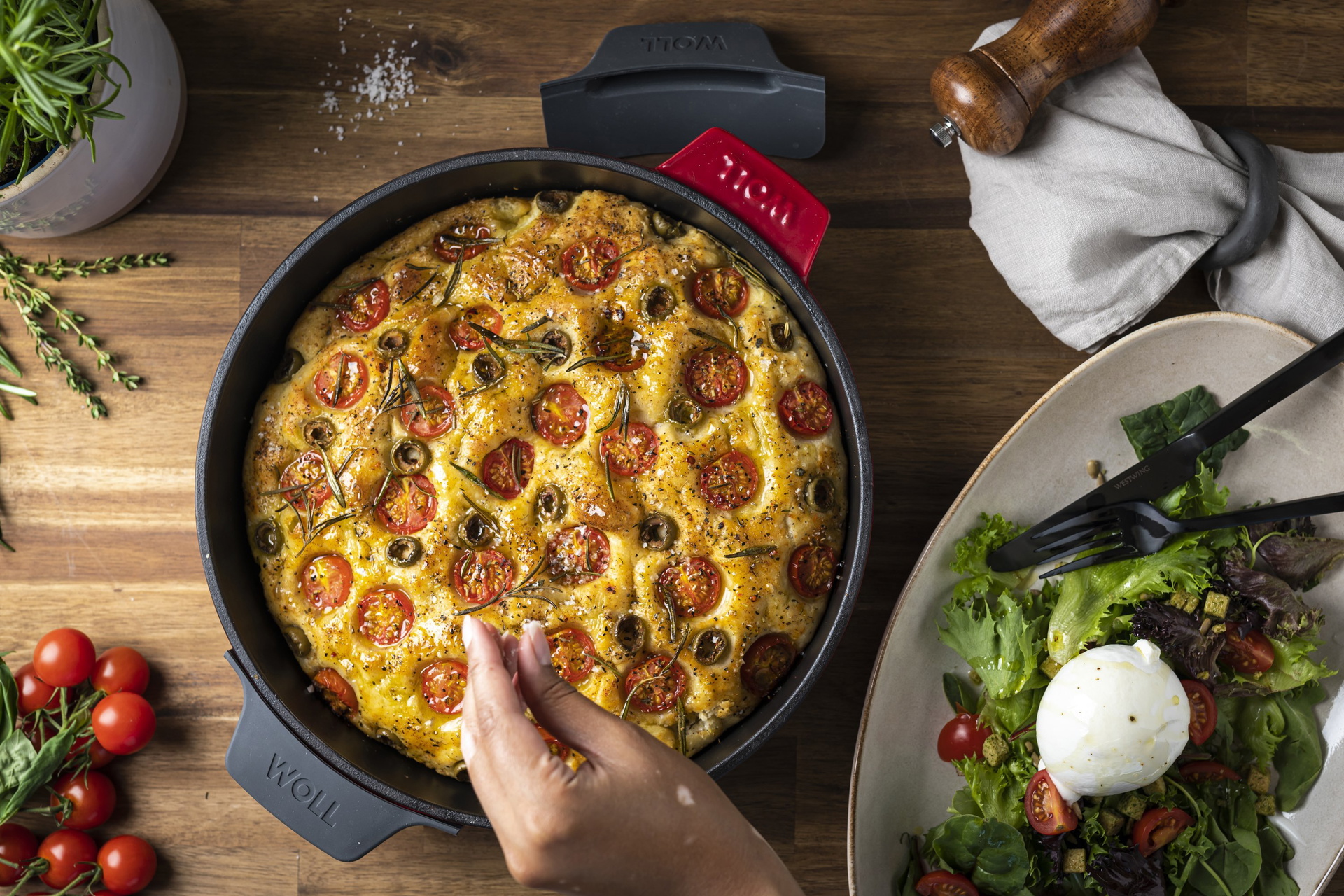 In der runden Kasserolle der Serie „Iron” befindet sich ein frisch gebackenes Focaccia, das mit Rosmarin und Meersalz garniert ist. Daneben stehen ein frischer Salat mit Burrata, frischen Kirschtomaten, ein Küchentuch, eine Mühle und frische Kräuter im To