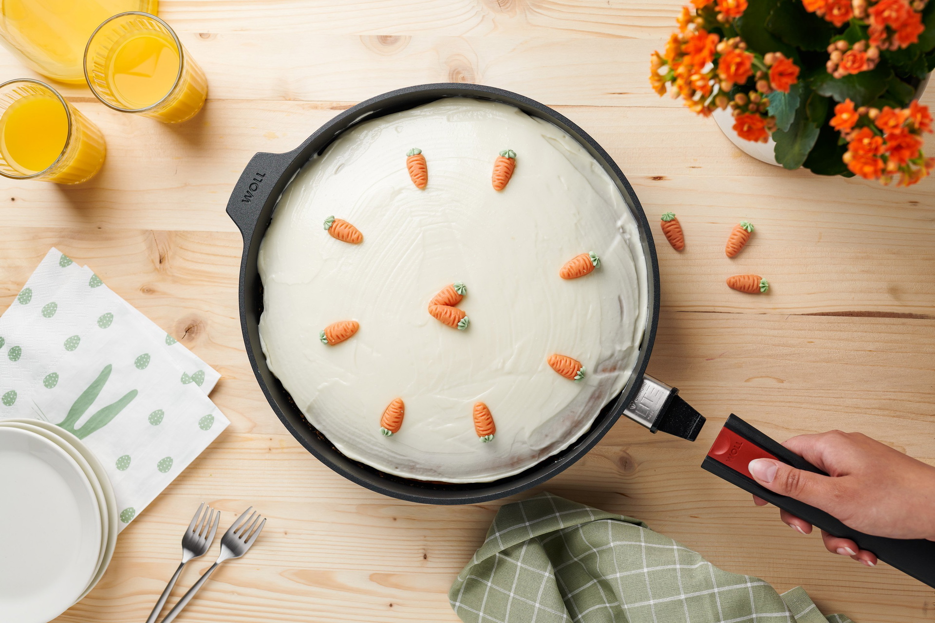 In einer runden Diamond-Lite-Pfanne mit abgenommem Stiel befindet sich frisch gebackener Karottenkuchen, der mit Marzipan-Karotten dekoriert ist. Daneben stehen Gläser mit Orangensaft und Teller mit Besteck.