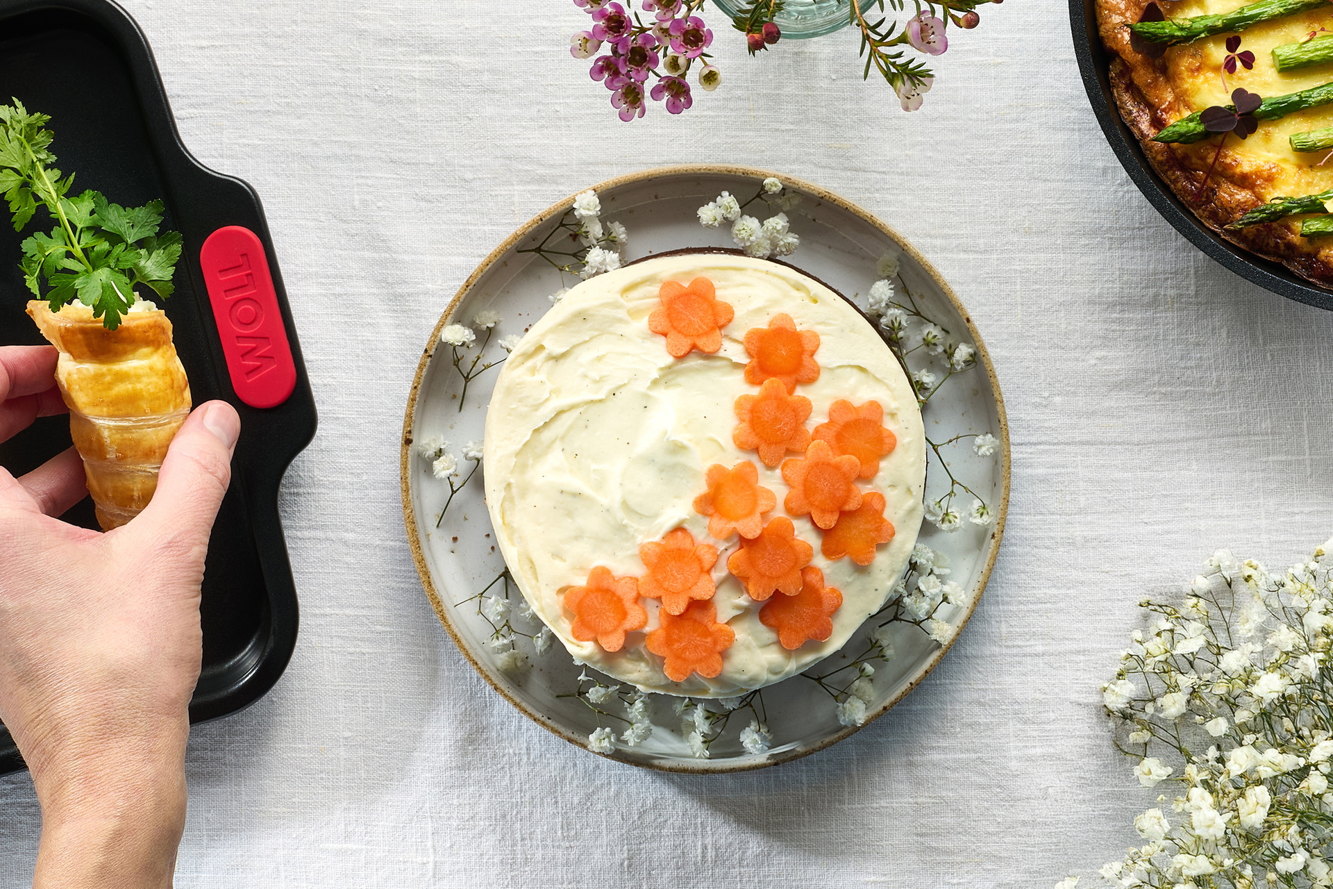 Auf einem Teller steht ein frisch gebackener Karottenkuchen mit Frischkäseglasur und Karottenblumen. Links daneben befindet sich ein Backblech aus der Serie „Let's Bake!” mit frisch gebackenem Blätterteig Karotten, rechts daneben ein weiterer frisch gebac