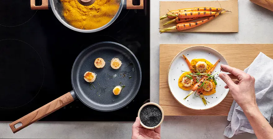 Die Jakobsmuscheln wurden in einer heißen, runden Pfanne der Serie Eco Logic QXR angebraten. Auf einem Teller, der neben dem Herd bereitsteht, werden sie auf einem Bett aus Karottenpüree serviert und mit einer halbierten, angebratenen Karotte garniert.