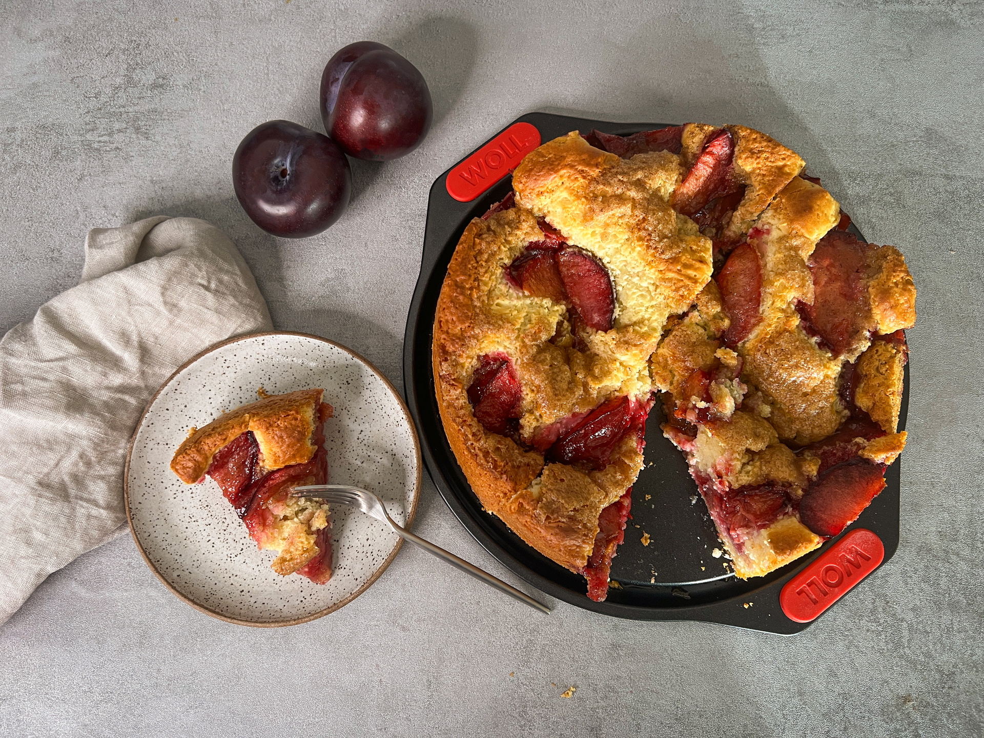 In einer runden Springform mit Schnellverriegelungen aus der Serie Let's Bake! befindet sich ein frisch gebackener Pflaumenkuchen mit Hefeteig und Zimt-Karamell-Topping. Daneben steht ein Teller mit einem Stück Kuchen, darüber sind zwei Pflaumen zu sehen.