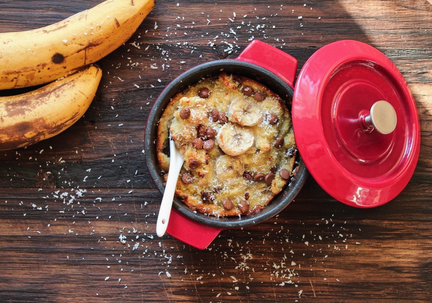 Auf der Arbeitsplatte steht eine runde Mini-Kasserolle der Serie Iron. Der Deckel wurde abgenommen und liegt schräg angelehnt daran. In der Kasserolle befindet sich frisch gebackener Bananenkuchen. Daneben liegen zwei Bananen.