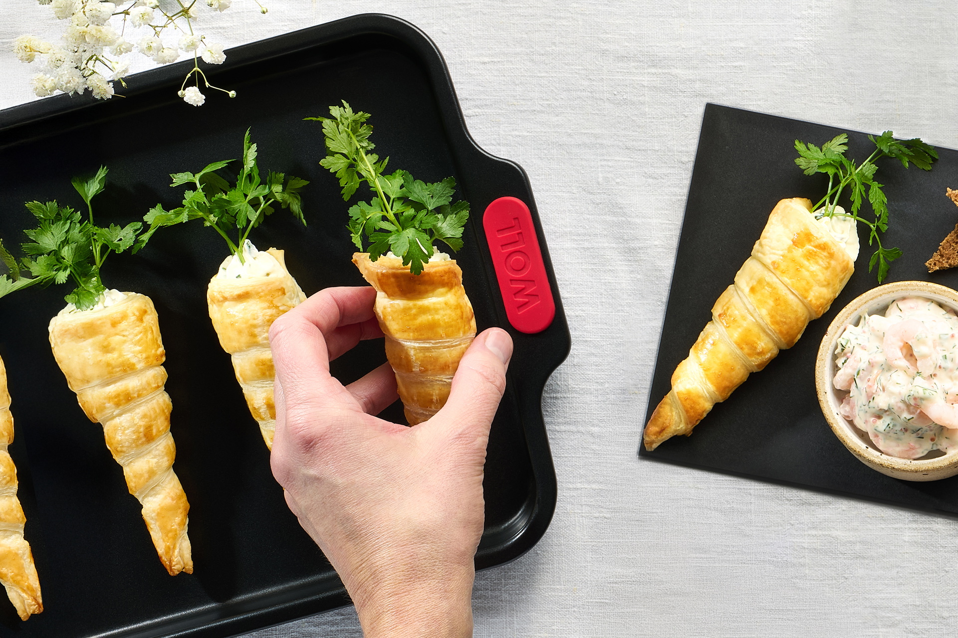 Auf einem Tisch steht ein Backblech der Serie „Let's Bake!”, auf dem frisch gebackene Blätterteig-Karotten mit Frischkäsefüllung liegen. Daneben liegt eine weitere Blätterteig-Karotte auf einem Schneidebrett, auf dem eine Schüssel mit Garnelen steht.