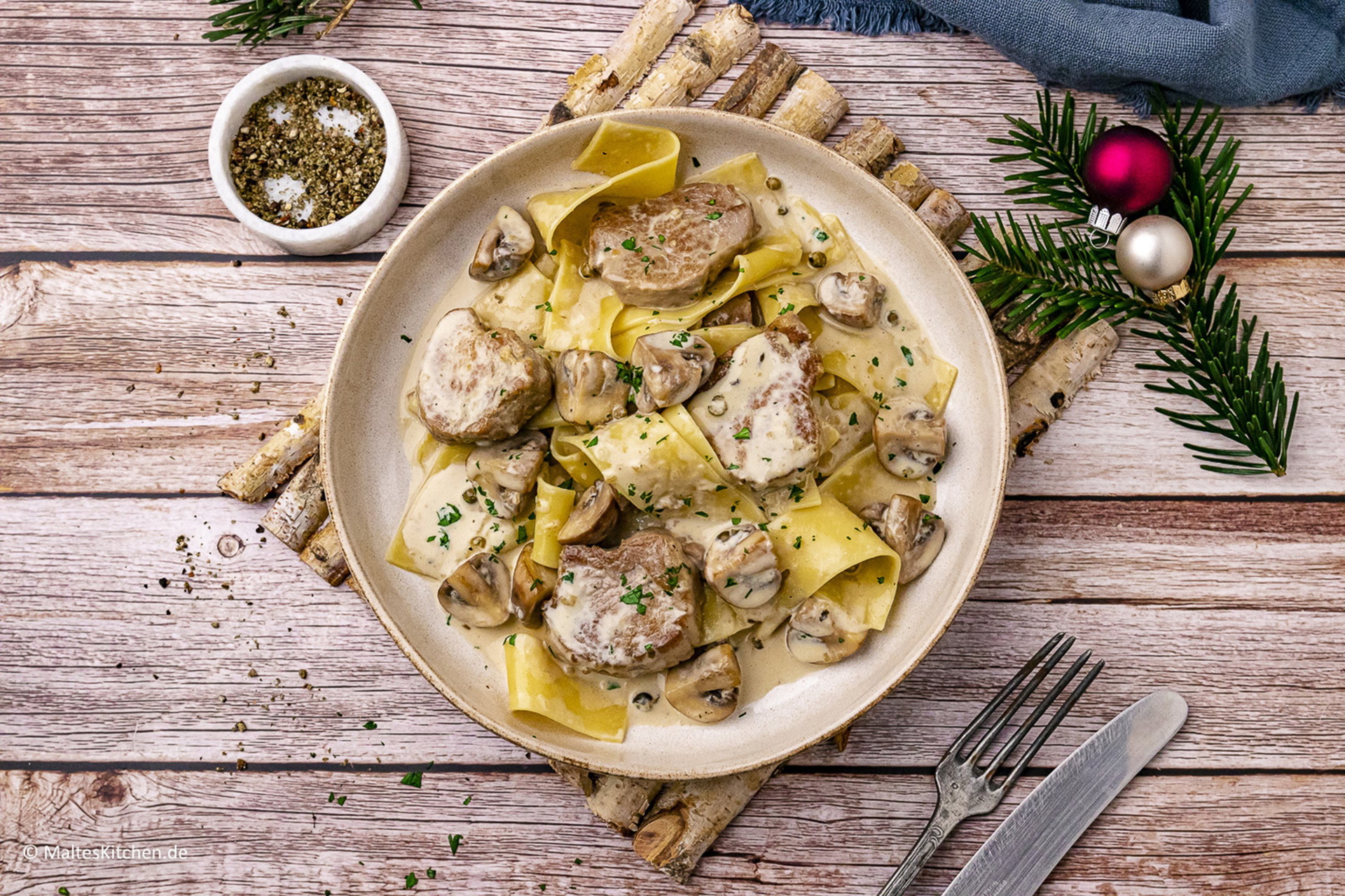 Auf einem Teller befindet sich frisch zubereitete Schweinelenden in creamiger Pilzrahmsoße. Daneben liegen Besteck, Gewürze und Weihnachtsdeko.
