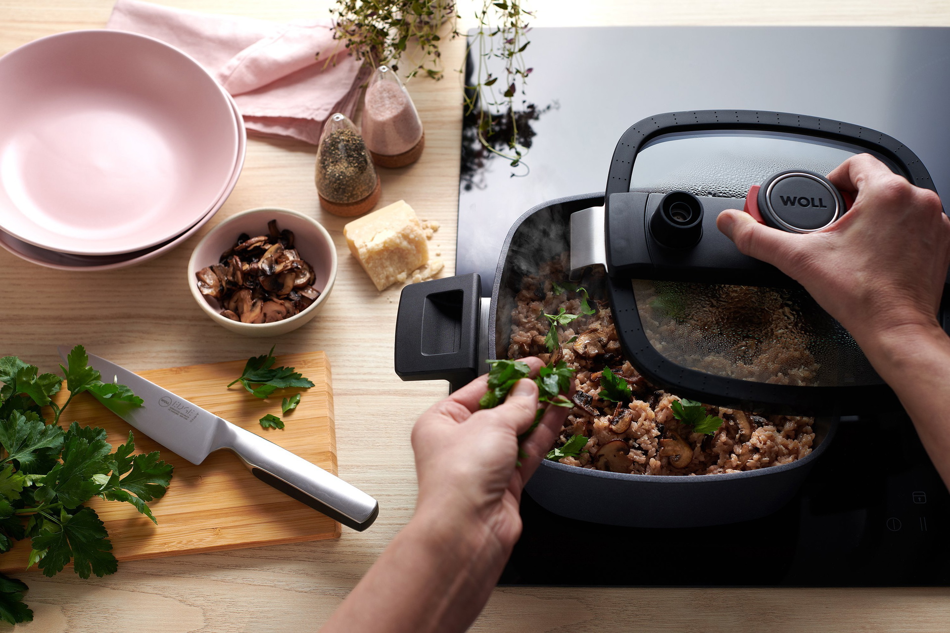 In einem heiße, viereckigen Niederdrucktopf der Serie Diamond Active Lite wird ein dampfendes Reisgericht zubereitet. Der Glasdeckel wurde von einer Hand entfernt. In der Nähe stehen eine kleine Schüssel mit fertigen Pilzen, ein Holzbrett mit einem Messer