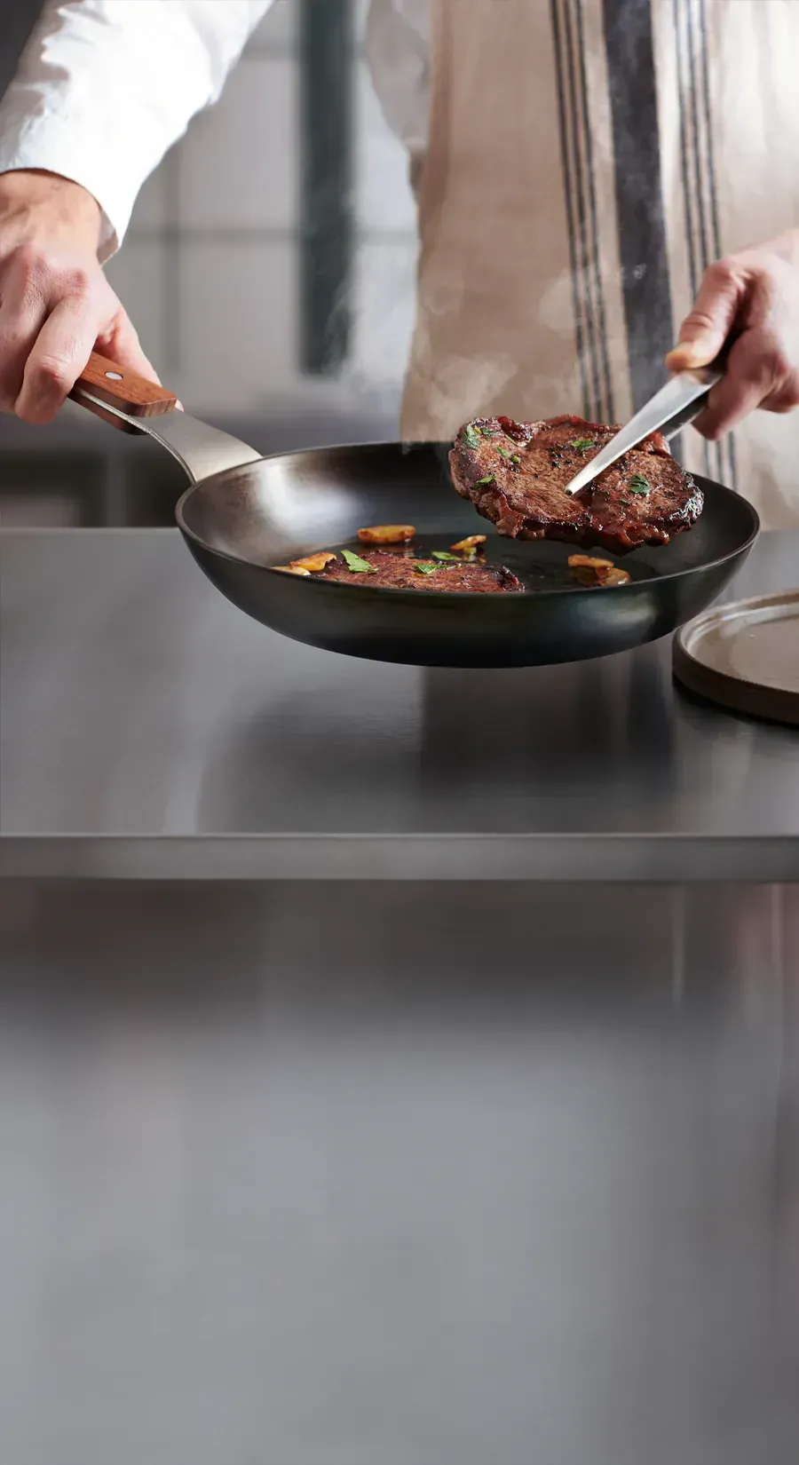 In einer heißen runden Carbon-Steel-Pfanne befinden sich zwei frisch, angebratene Steaks. Eins davon wird mit einer Zange herausgenommen, um dieses auf den daneben stehenden Teller zu platzieren.