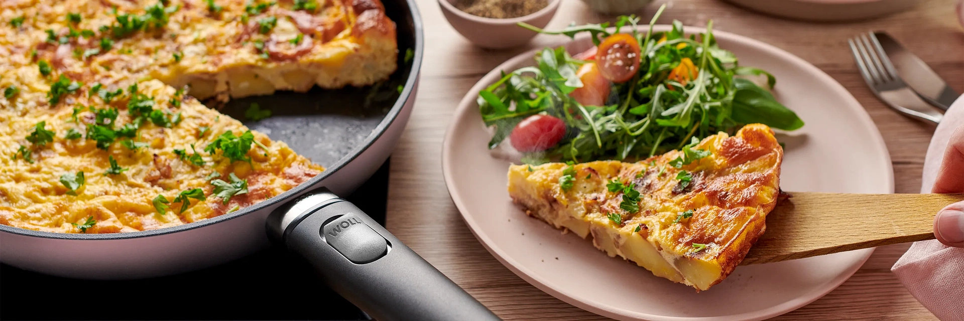 In einer runden Pfanne der Serie Ecolour mit abnehmbarem Stiel befindet sich eine frisch zubereitete spanische Tortilla. Ein Teil davon wird zusammen mit frischem Salat auf einem Teller serviert.