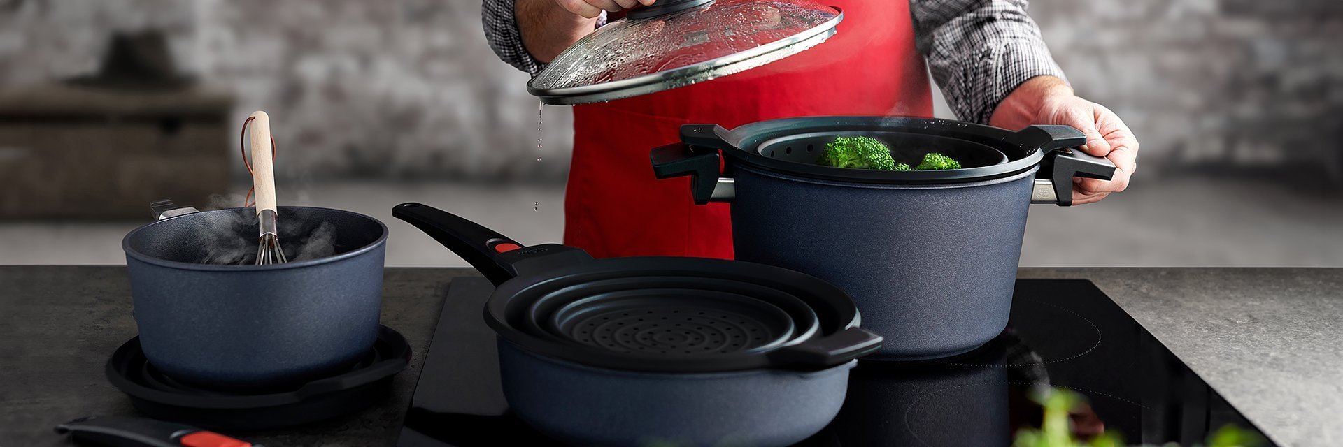Auf dem Herd befinden sich eine runde Pfanne und ein runder Topf der Serie Diamond Lite, bei dem der Glasdeckel abgenommen wurde. Beide verwenden die Silikoneinsätze der Serie Steam it. Im Topf wird Gemüse gedünstet, während der Silikoneinsatz der Pfanne