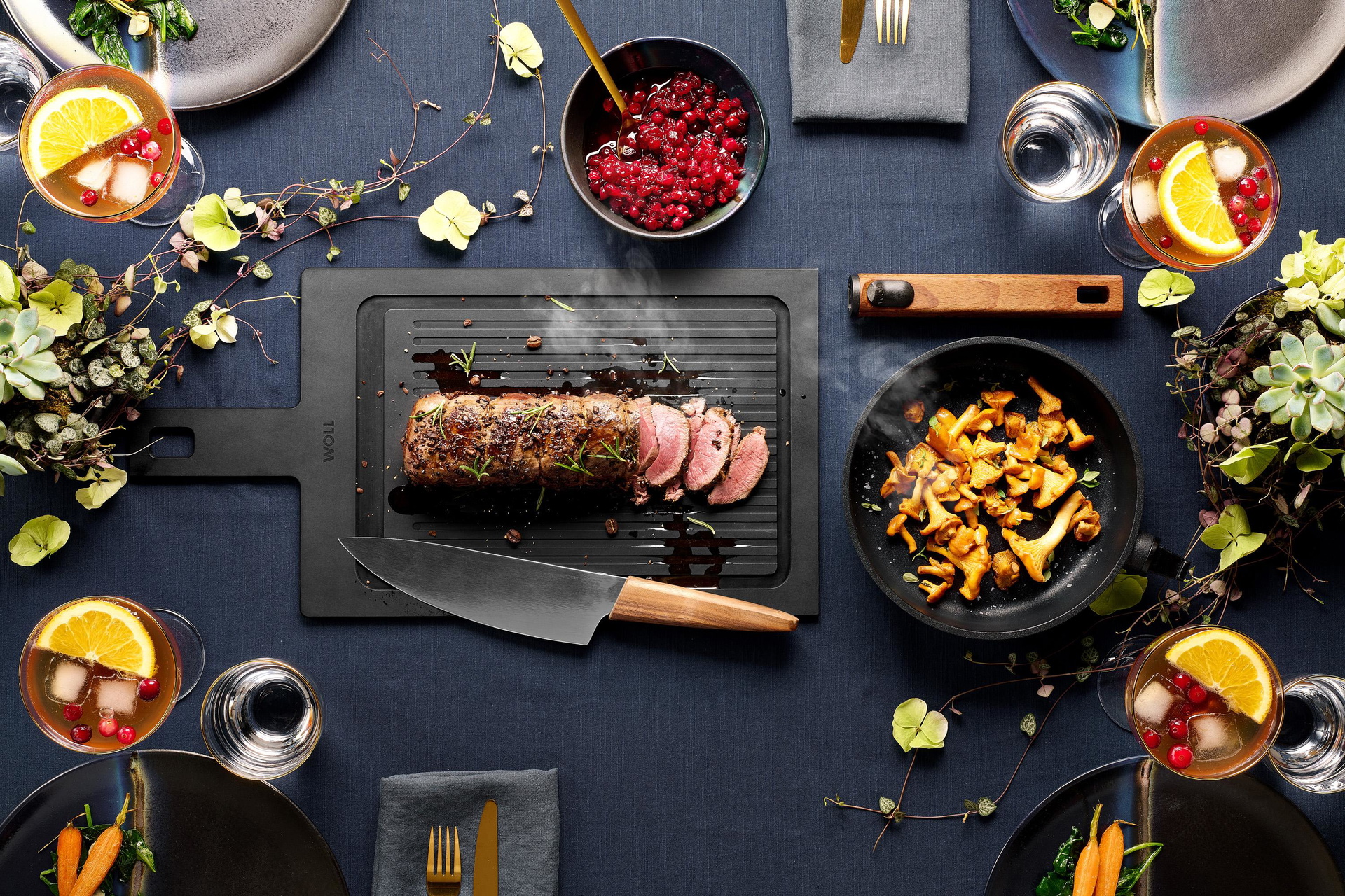 Auf einem Schneidebrett der Serie „Cut & Serve“ mit Rillen und Saftrille liegt ein dampfendes Stück Rehrücken mit Kaffee-Crunch, von dem bereits einige Scheiben geschnitten wurden. In der runden, heißen Eco-Logic-QXR-Pfanne daneben befinden sich frisch an