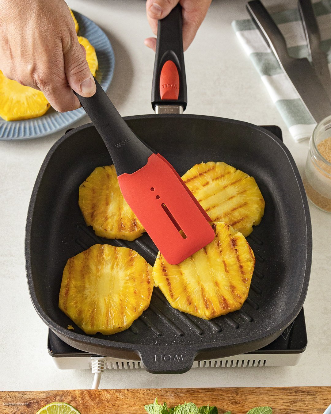 In einer viereckigen Titan Best Pfanne werden Ananasscheiben angebraten und mit einer Cock it Küchenzange gewendet.