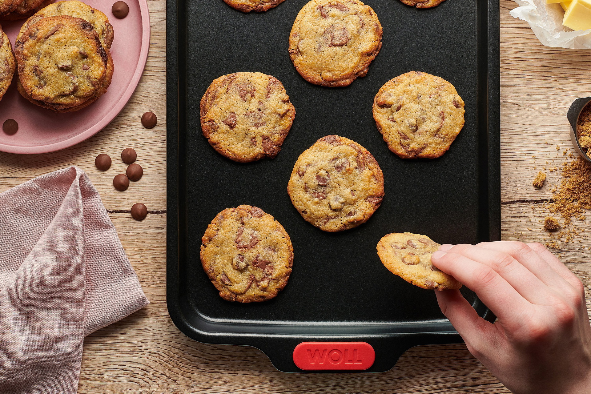 Auf einem rechteckigen Backblech der Serie Let's Bake! liegen frisch gebackene Cookies mit Schokoladestückchen. Ein Cookie wird vom Blech genommen. Auf dem Teller daneben liegen weitere gebackene Cookies. Rechts vom Blech liegen weitere Zutaten.