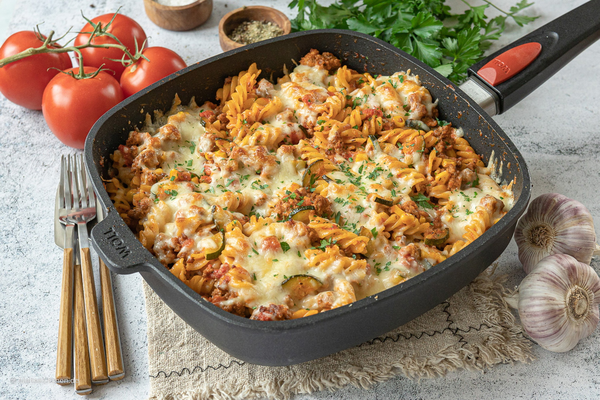 In einer viereckigen Titan-Best-Pfanne mit abnehmbarem Stiel befindet sich ein frisch zubereiteter Nudelauflauf mit Hackfleisch und Zucchini. Daneben liegen Besteck, ganze Tomaten, Knoblauchknollen, Gewürze und frischer Petersilie.
