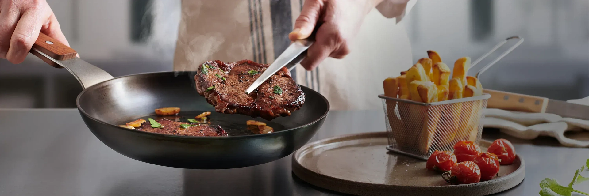 In einer heißen runden Carbon Steel Pfanne mit festem Holzstiel befinden sich zwei frisch, angebratene Steaks. Eins davon wird mit einer Zange herausgenommen, um dieses auf den daneben stehenden Teller zu platzieren. Auf diesem Teller steht ein kleiner Ko