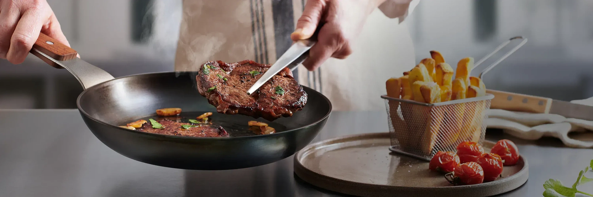 In einer heißen runden Carbon Steel Pfanne mit festem Holzstiel befinden sich zwei frisch, angebratene Steaks. Eins davon wird mit einer Zange herausgenommen, um dieses auf den daneben stehenden Teller zu platzieren. Auf diesem Teller steht ein kleiner Ko