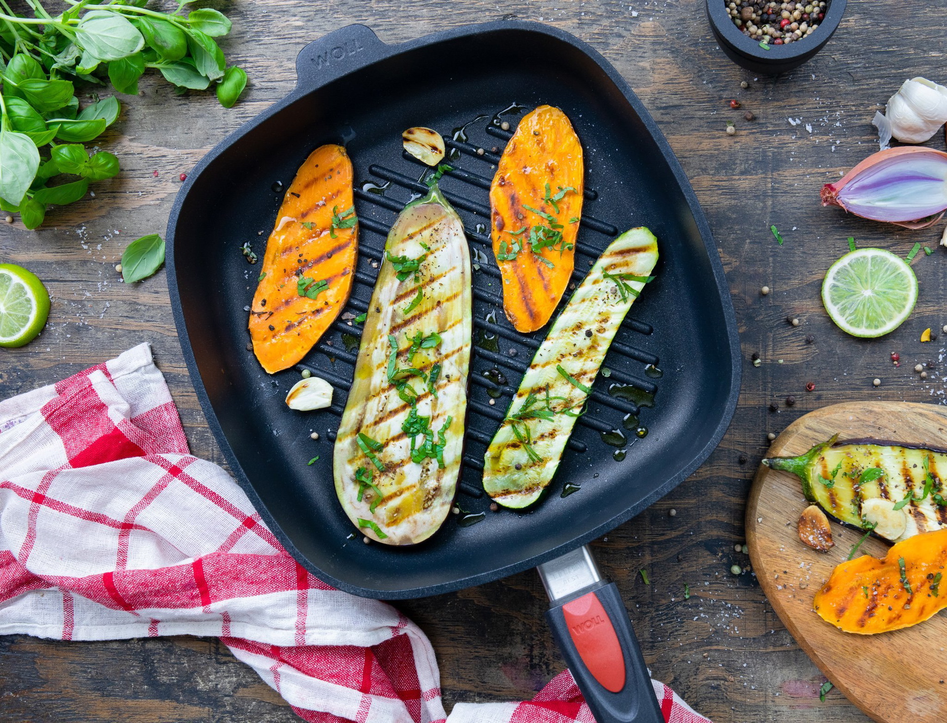 In einer viereckigen, heißen Titan-Beste-Grillpfanne mit Rillen und abnehmbarem Stiel. Darin befindet sich frisch zubereitetes, dampfendes Grillgemüse. Auf einem Holzbrett liegen gegrilltes Gemüse und außenrum weitere Zutaten.