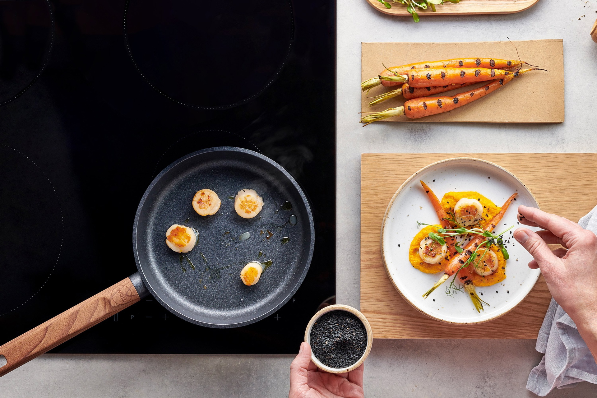 Die Jakobsmuscheln wurden in einer runden, heißen Pfanne der Serie „Eco Logic QXR” mit festem Holzstiel angebraten. Auf einem neben dem Herd bereitstehenden Teller wurden die Jakobsmuscheln mit Karottenpüree, gerösteten Möhren, frischen Sprossen und schwa