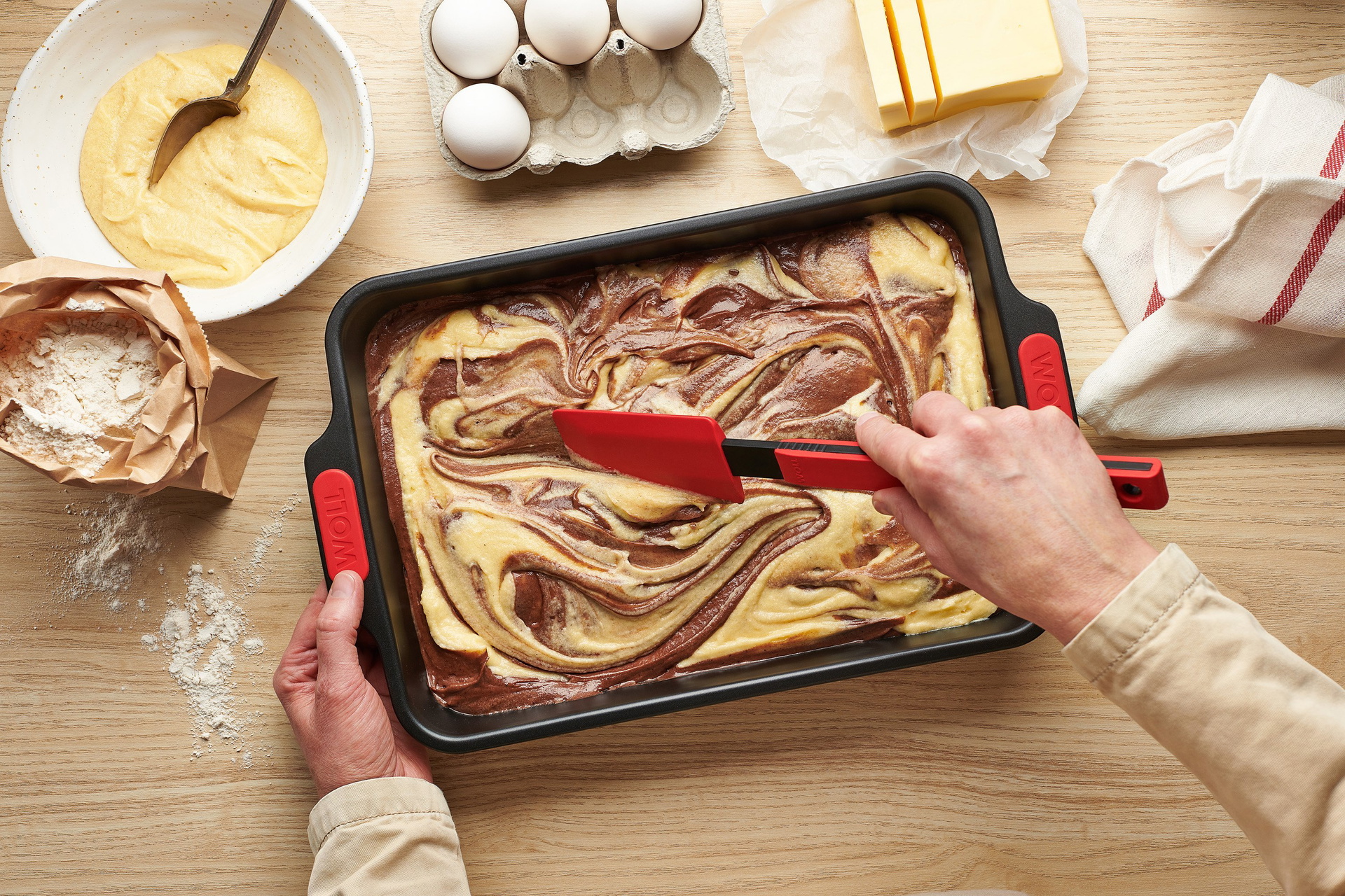 In der Backform der Serie Let’s Bake! befindet sich roher Marmorkuchenteig, der mit einem Teigschaber der Serie Cook i” verteilt wird. Daneben sind eine offene Packung Mehl, ein Küchentuch sowie eine Schale mit Teig zu sehen.