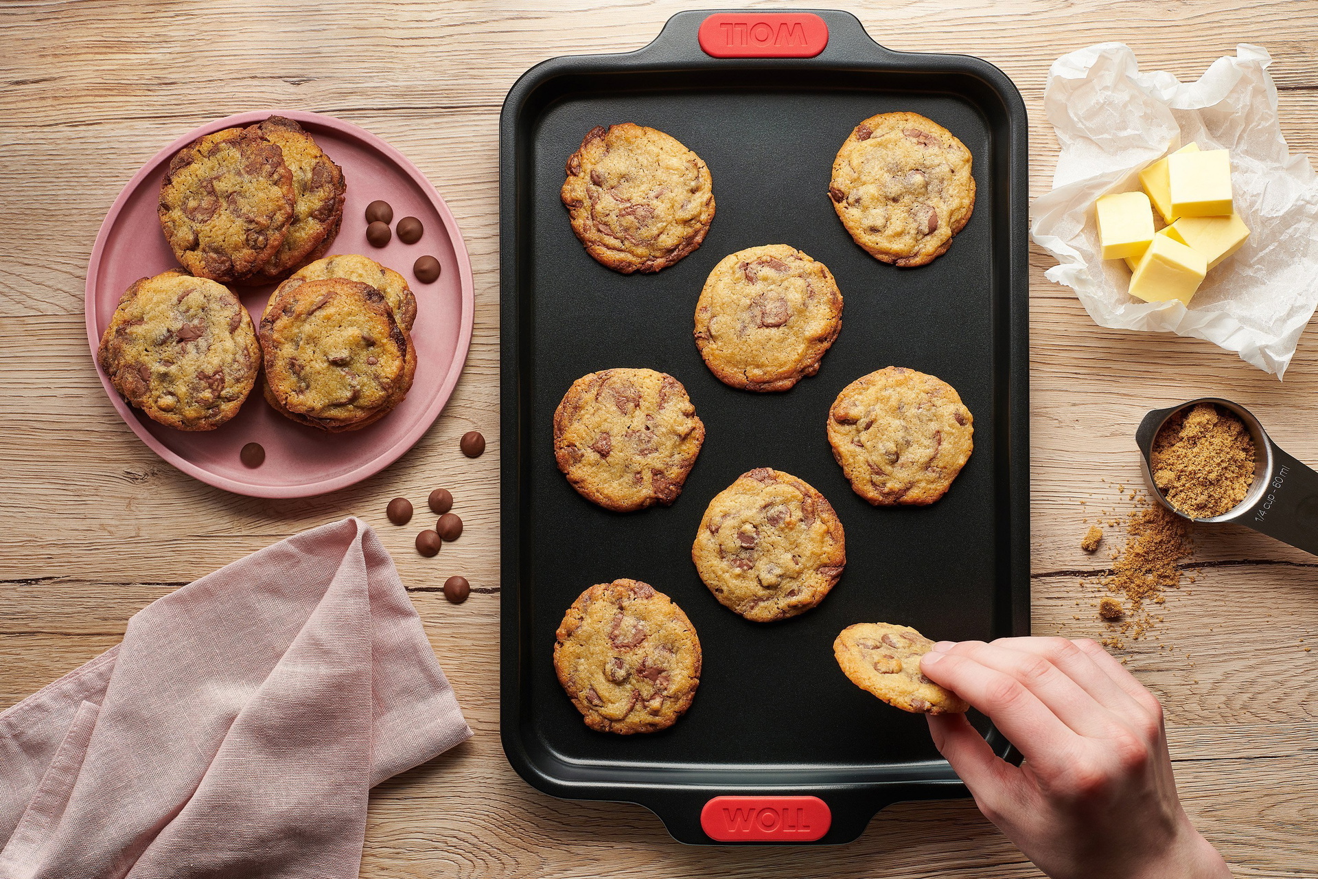 Auf einem rechteckigen Let's bake! Backblech liegen frisch gebackene Cookies mit Schokoladestückchen. Ein Cookie wird vom Blech genommen.