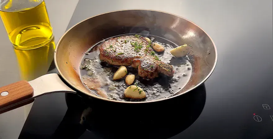 In einer runden, heißen Carbon-Steel-Pfanne mit Antihaftbeschichtung wird ein Stück Fleisch mit Kartoffeln angebraten. Daneben steht eine Flasche mit neutralem Öl.