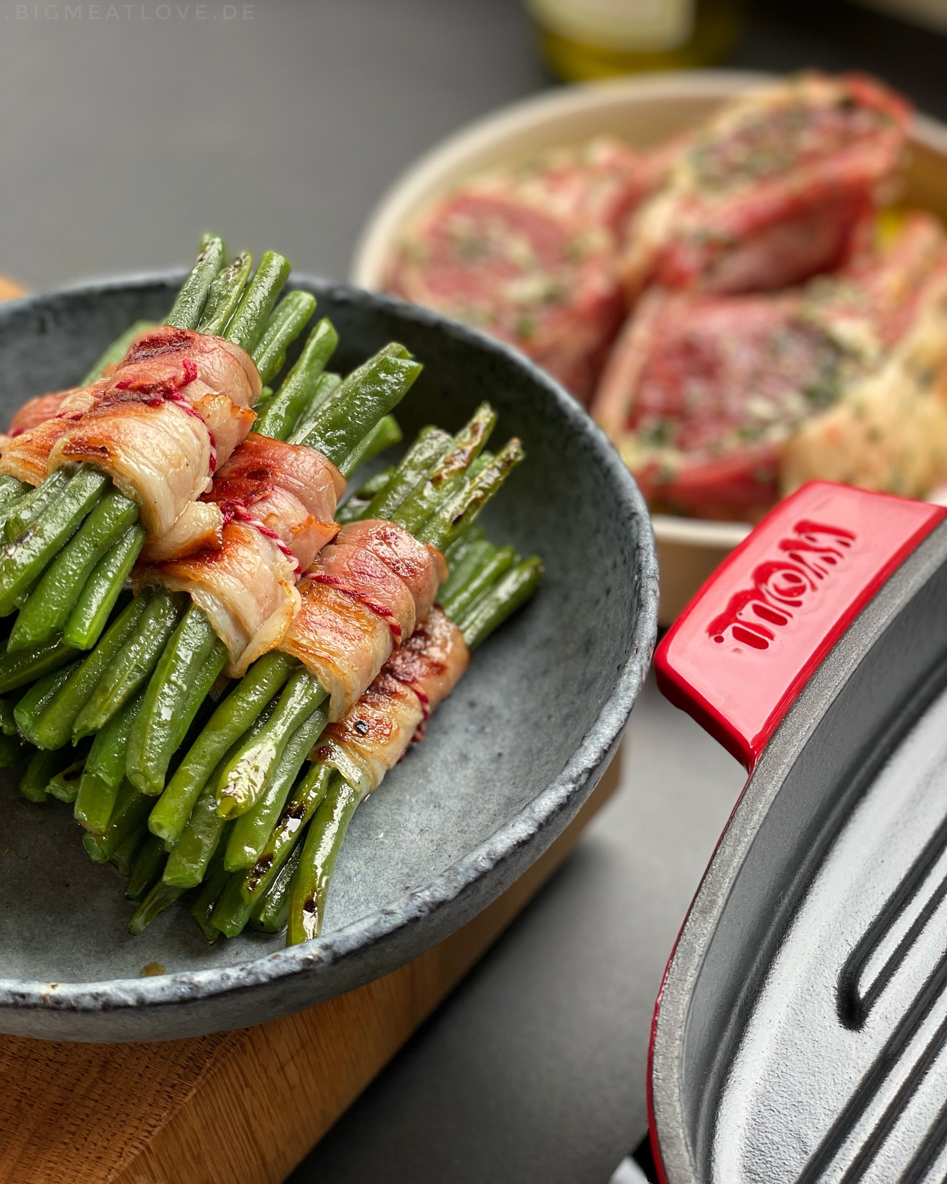 image1 Auf dem Herd steht eine viereckige, rote Iron Grillpfanne mit Rillen. In einer Schüssel liegen angebratene Bohnen im Speckmantel, dahinter mariniertes Fleisch.