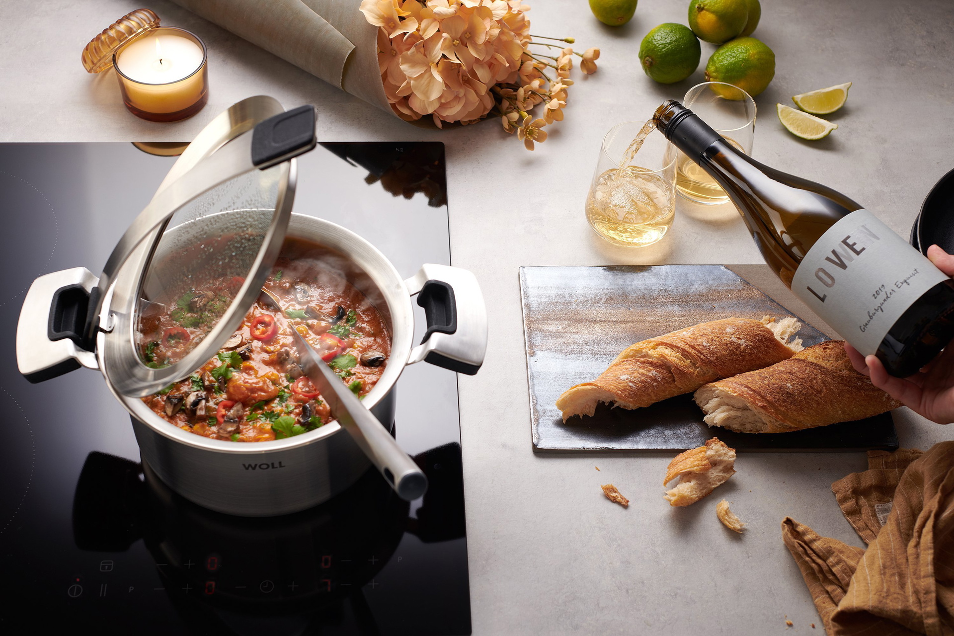 In einem runden Topf der Serie „Concept” befindet sich frisch gekochtes Pilz-Curry mit Spinat und Kichererbsen. Der Deckel ist in der Griffhalterung eingerastet. Auf der Arbeitsplatte liegen frisches Baguette und Weißwein wird in Gläser eingeschenkt.