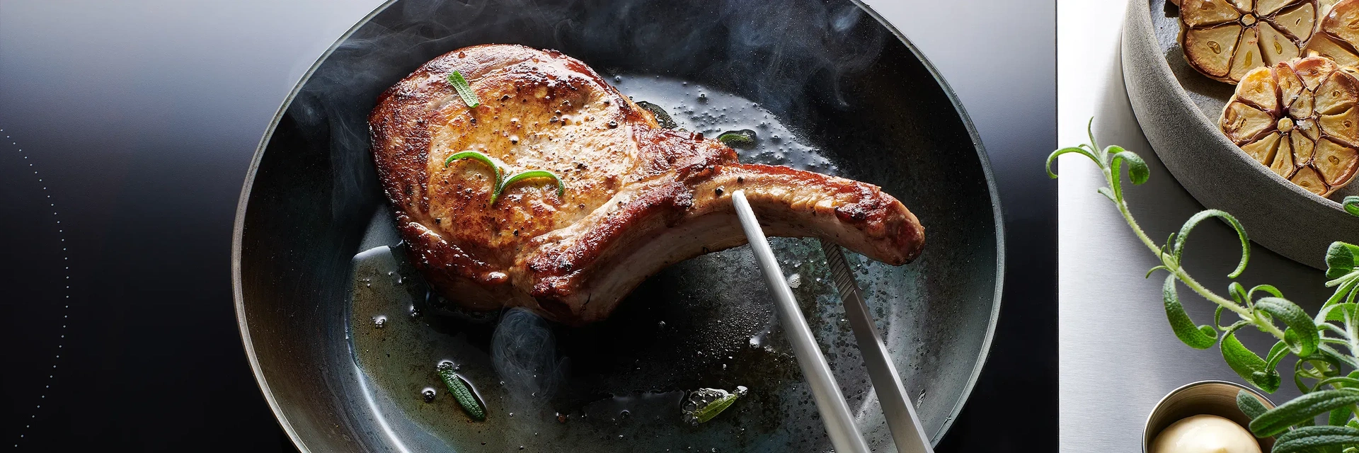 In einer Carbon-Steel-Pfanne mit eingebrannter Antihaftbeschichtung wird ein Stück Fleisch angebraten. Dieses wird mit einer Zange gewendet. Auf der daneben stehenden Arbeitsplatte steht eine Schale mit angebratenen, halbierten Knoblauchknollen.