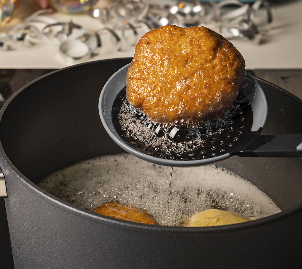 Mit einer grauen Cock it Schöpfkelle wird ein ausbackener Fastnachtkrapfen aus dem Topf entnommen und über dem Topf abgetropft.