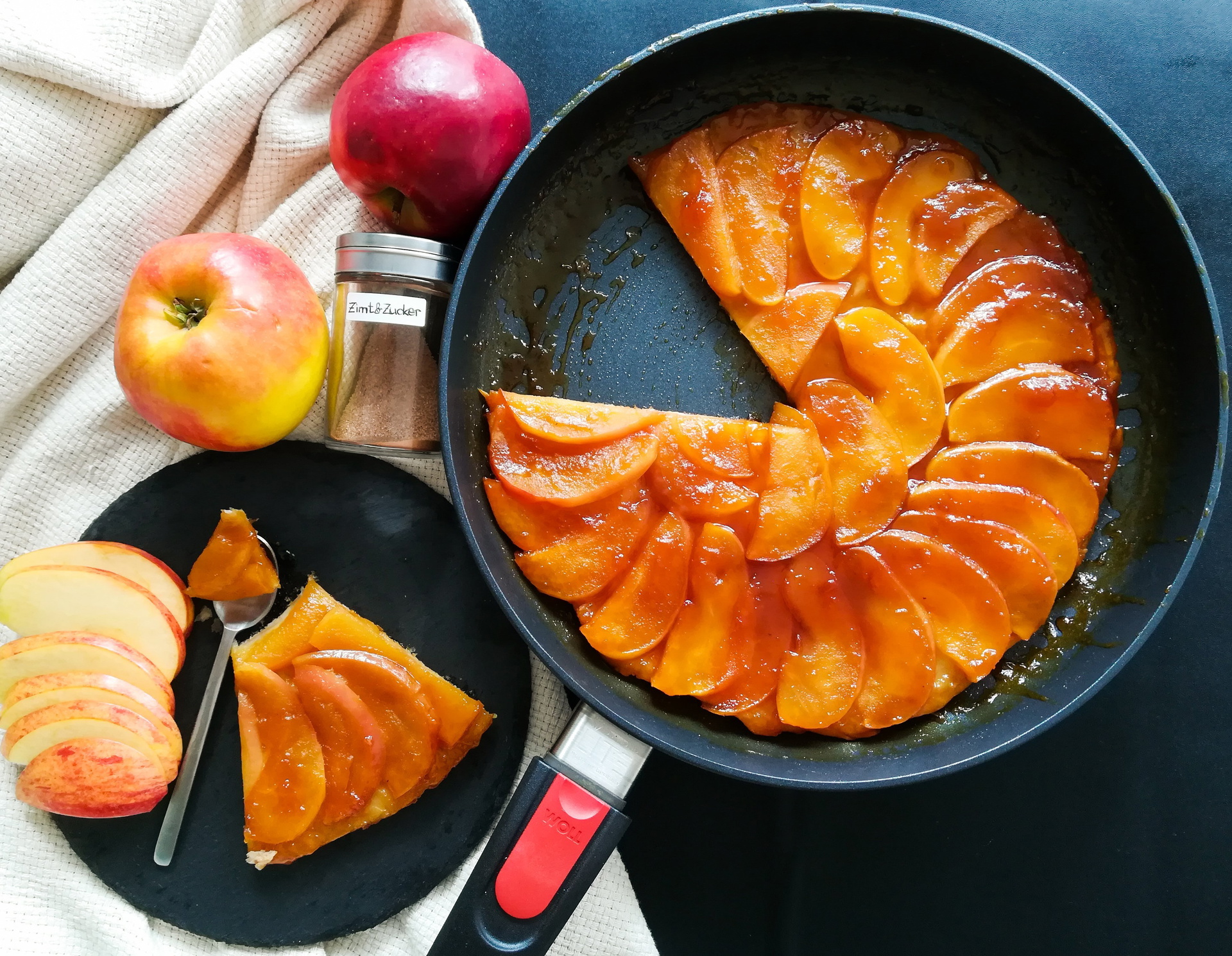 In einer runden Diamond-Lite-Pfanne mit abnehmbarem Stiel befindet sich eine frisch zubereitete, warme Apfeltarte. Ein Stück wurde entnommen und auf einem daneben platzierten Teller angerichtet. Auf dem Teller liegen außerdem Apfelspalten, darüber befinde
