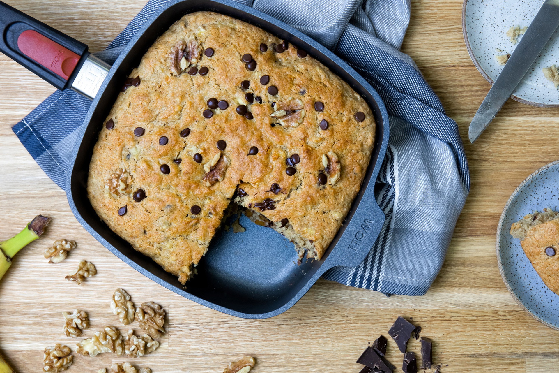 Auf einem Tisch steht eine viereckige Diamond-Lite-Pfanne mit abnehmbarem Stiel, in der frisch gebackenes Bananenbrot gebackenen wurde. Ein Stück wurde entnommen und auf dem danebenstehenden Teller angerichtet. Daneben steht ein weiterer Teller und außen