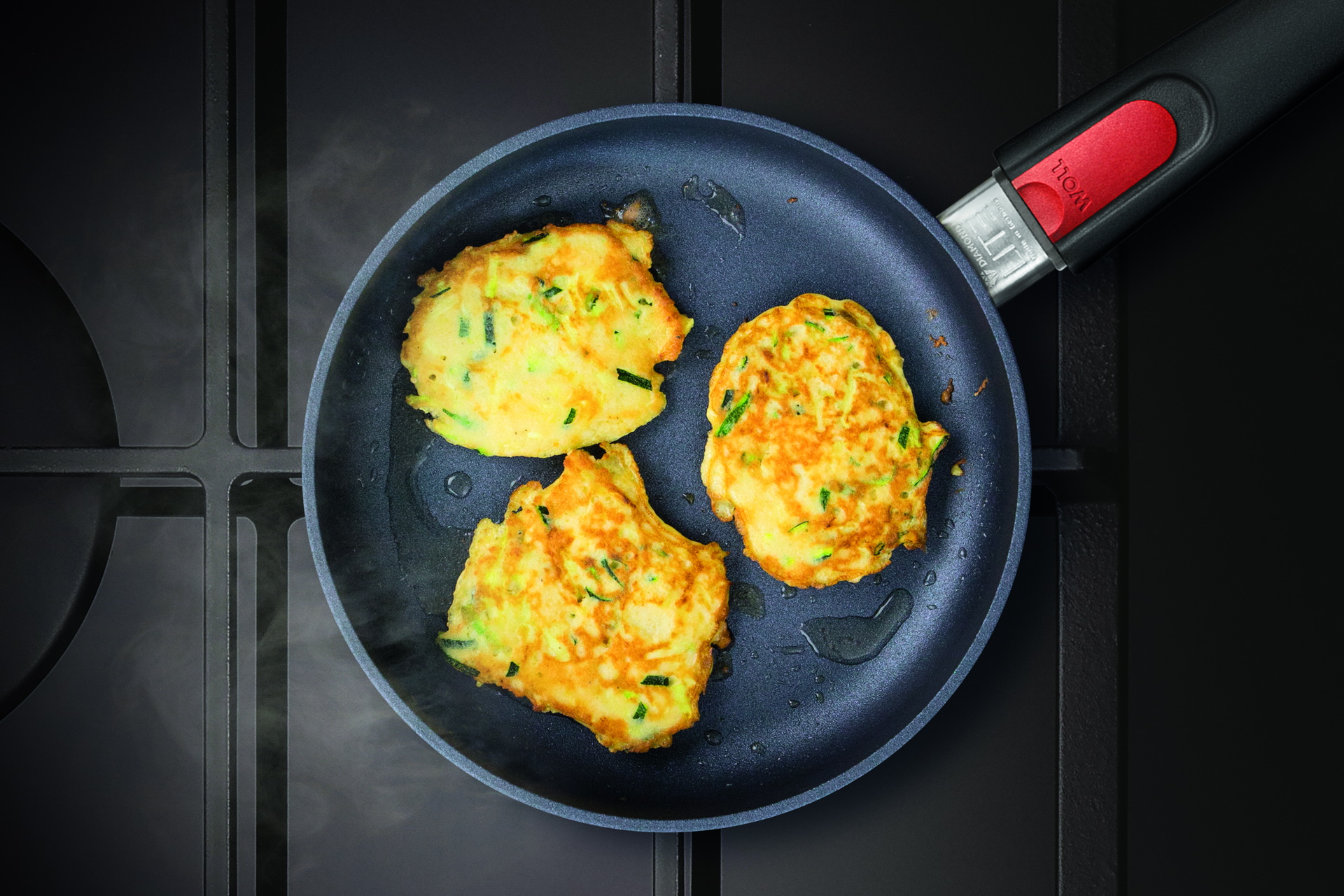 Auf dem Herd steht eine runde, heiße Diamond-Lite-Pfanne mit abnehmbarem Stiel, in der sich frisch zubereitete, dampfende Zucchini Pancakes befinden.