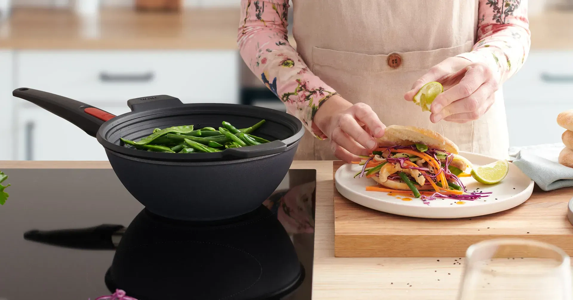 In dem runden Wok der Serie „Diamond Lite” wird mithilfe des Silikoneinsatzes „Steam it” Gemüse gedämpft. Daneben steht ein Teller mit frischem Essen, auf das etwas Limettensaft ausgepresst wird.