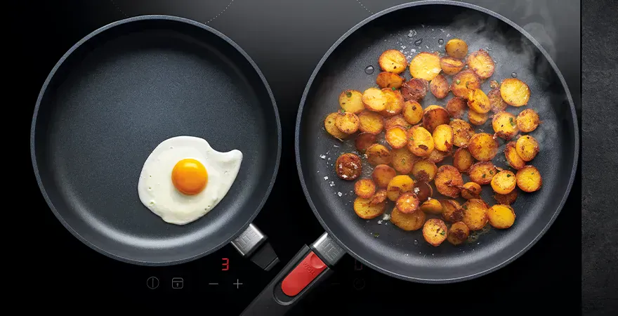 Auf dem Herd stehen zwei heiße, runde Diamond-Lite-Pfannen. In der linken Pfanne brät ein Spiegelei. Um sie sicher neben der rechten Pfanne platzieren zu können, wurde der Stiel entfernt. In der rechten Pfanne, die noch mit ihrem Stiel versehen ist, befin