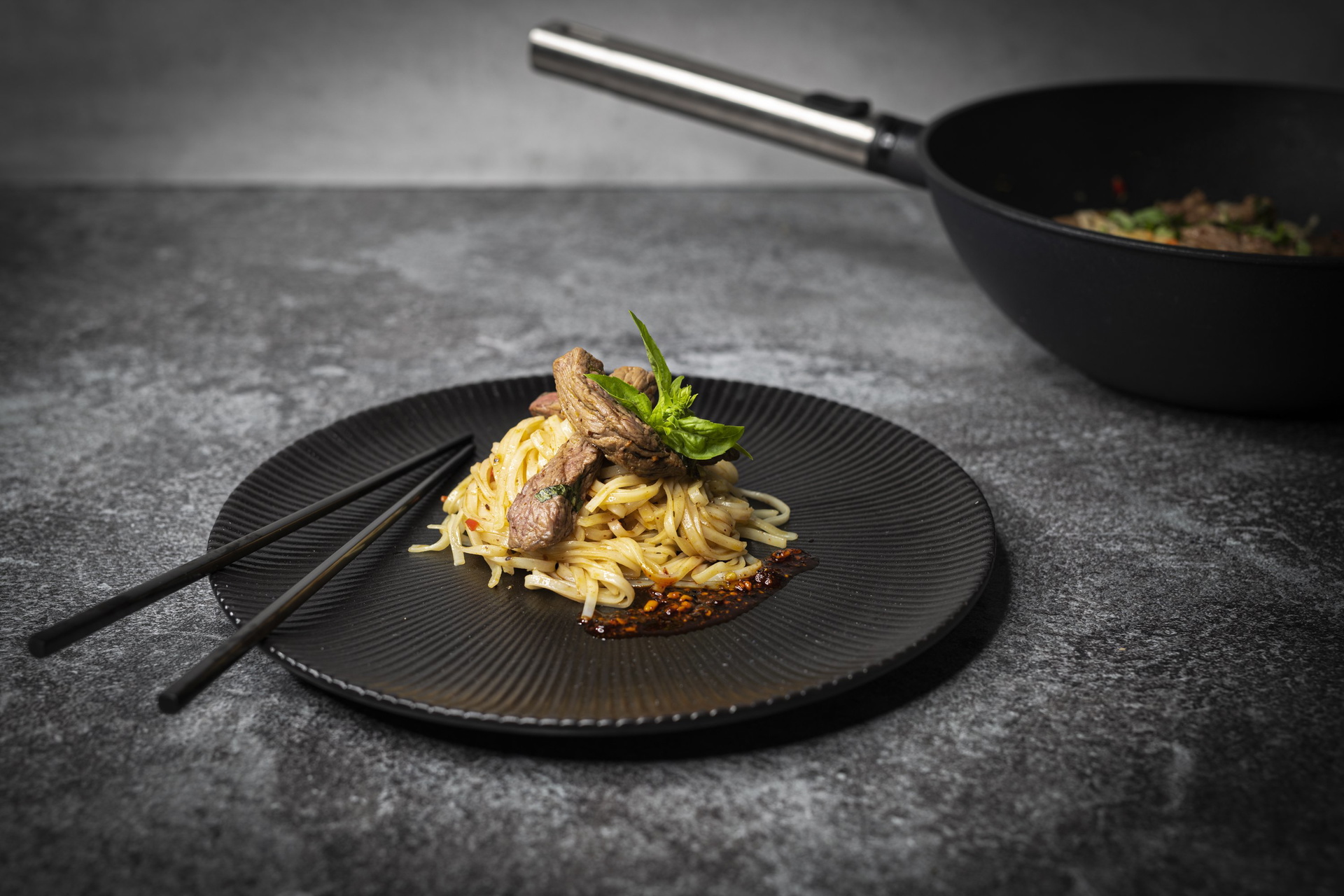 Auf der Arbeitsplatte steht eine Wok- und Rührpfanne der Serie Eco Logic QXR, in der Asia-Nudeln mit Steakstreifen zubereitet wurden. Eine Portion davon wurde auf einem daneben stehenden Teller angerichtet.