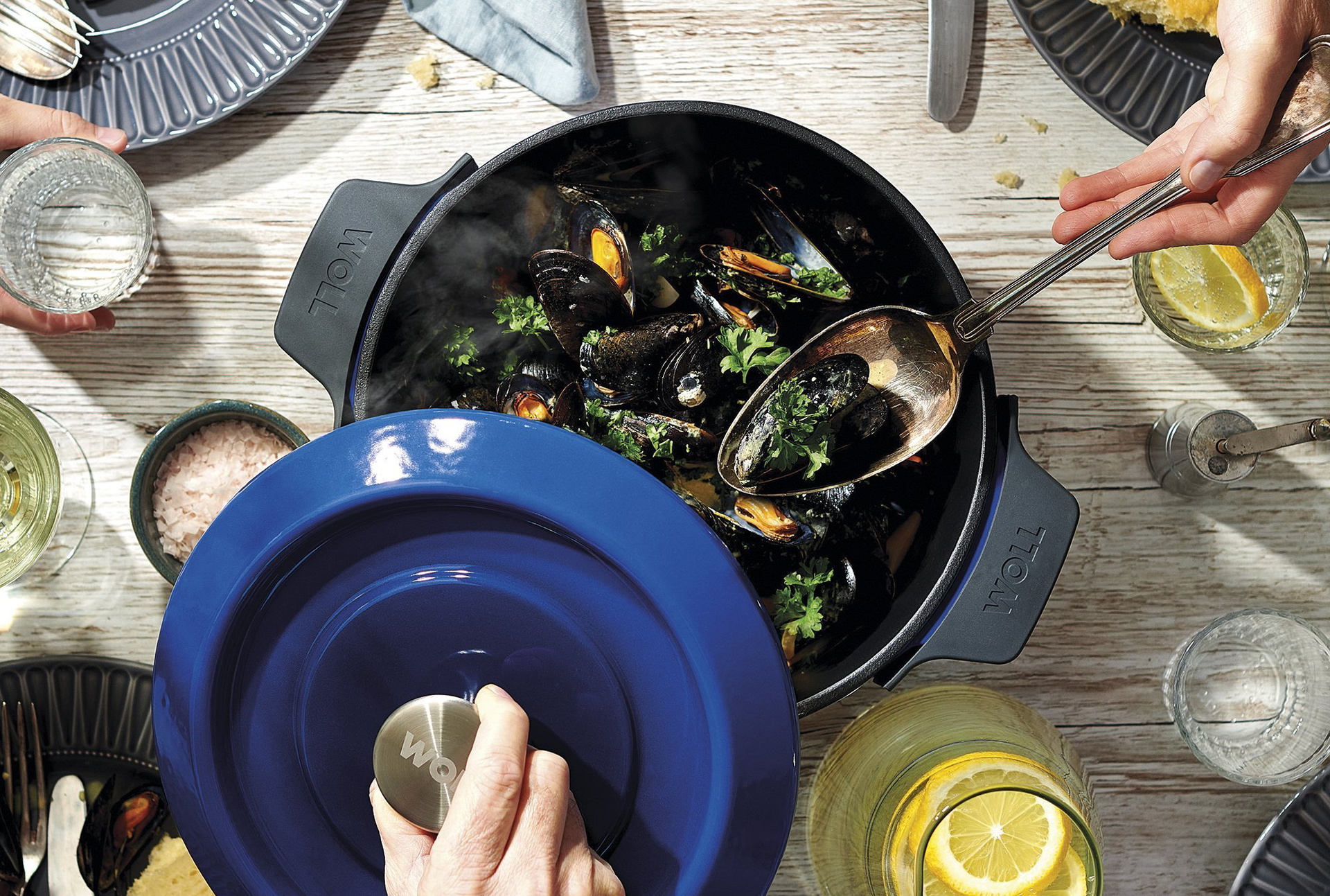 124CI-828CI_RGB Auf dem Tisch steht eine runde, blaue Iron-Kasserolle mit zubereiteten Muscheln. Der Deckel wird hochgehoben.