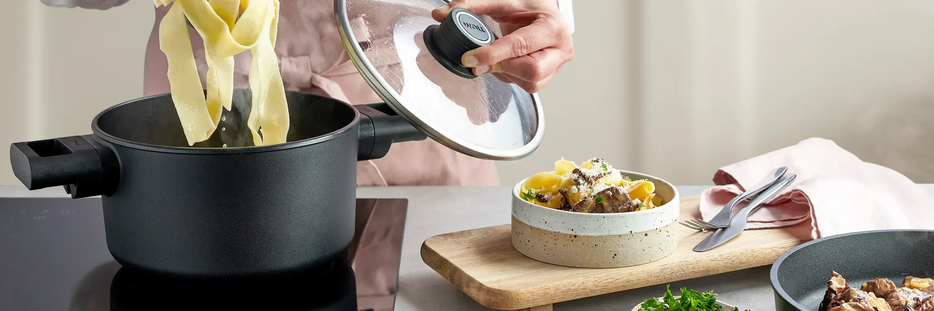 In einem runden Topf der Serie Eco Logic QXR werden die frisch gekochten Nudeln entnommen, während der Deckel mit einer Hand festgehalten wird. Rechts daneben steht auf einem Holzbrett ein fertig gemachtes Nudelgericht.