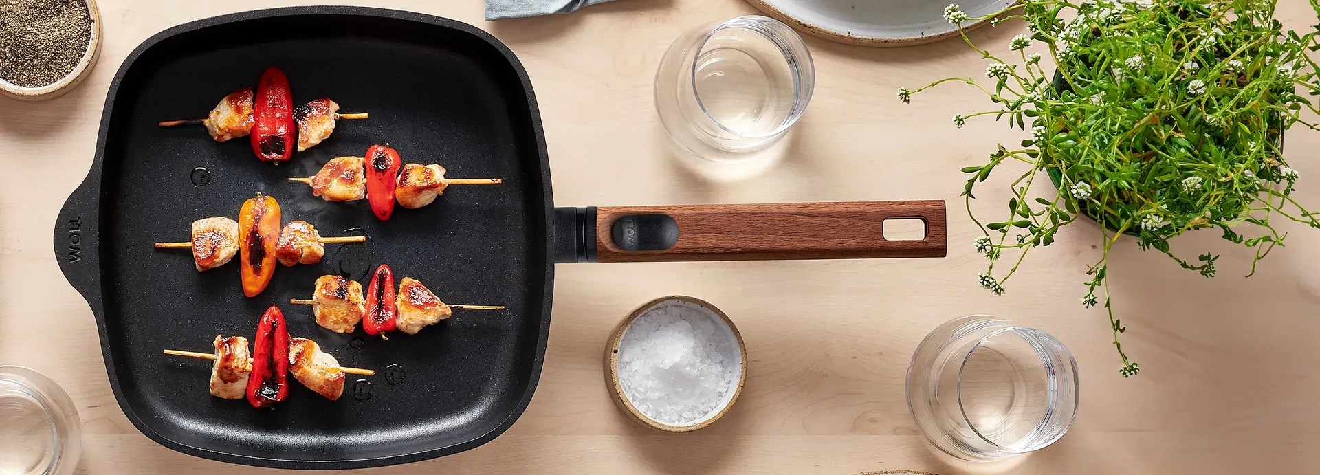 Auf dem Tisch steht eine viereckige Pfanne mit abnehmbarem Stiel aus der Serie Eco Logic QXR, die frisch angebratene Fleischspieße enthält. Drumherum stehen Teller, Gläser, Gewürze und eine Pflanze.