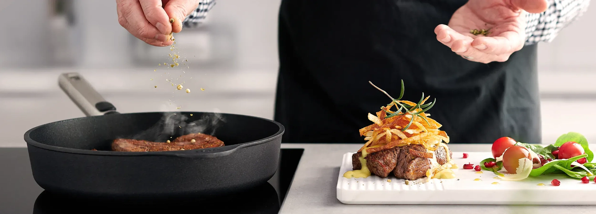 In einer heißen, runden Logic-Pfanne wird frisches Fleisch angebraten. Daneben wird ein weiteres Stück Fleisch mit gerösteten Zwiebeln und Soße angerichtet. Auf dem Brett liegt außerdem Salat.