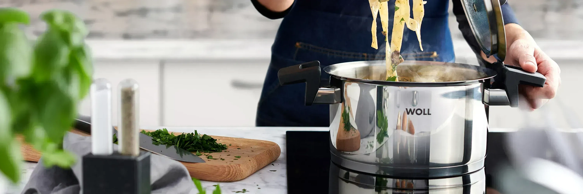 Im runden Edelstahltopf der Serie „Concept” befinden sich frisch zubereitete Bandnudeln. Der Deckel wurde abgenommen und in die dafür vorgesehene Halterung am Griff gesteckt. Daneben liegt ein Schneidebrett mit frischen Kräutern und einem Messer darauf.