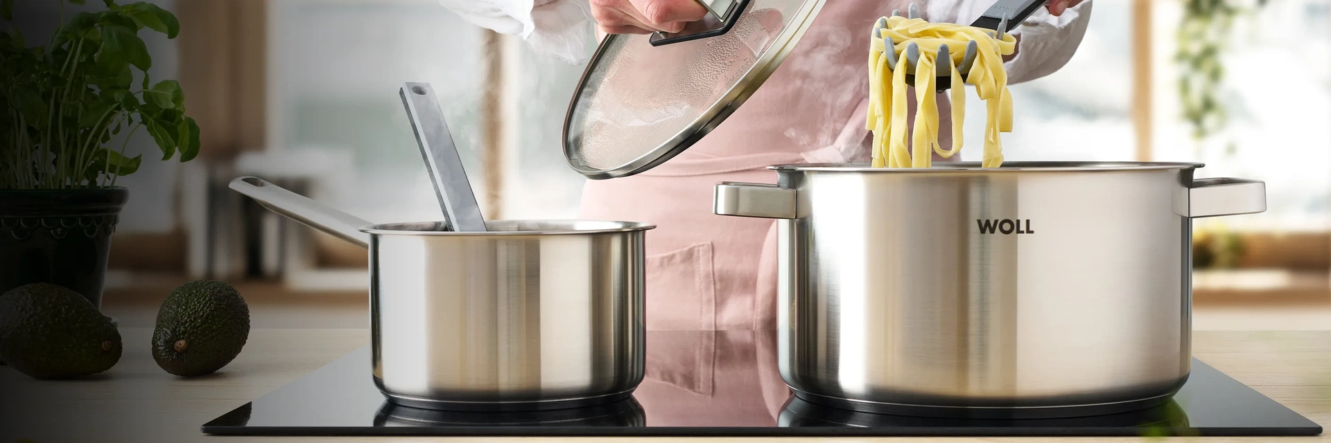 In einem kleinen, runden Topf dampft ein Gericht. Daneben werden die frisch gekochten Nudeln mit einem Pastalöffel aus dem Topf genommen. Hierzu wird der Glasdeckel angehoben. Auf der Arbeitsplatte liegen Avocado und Kräuter.