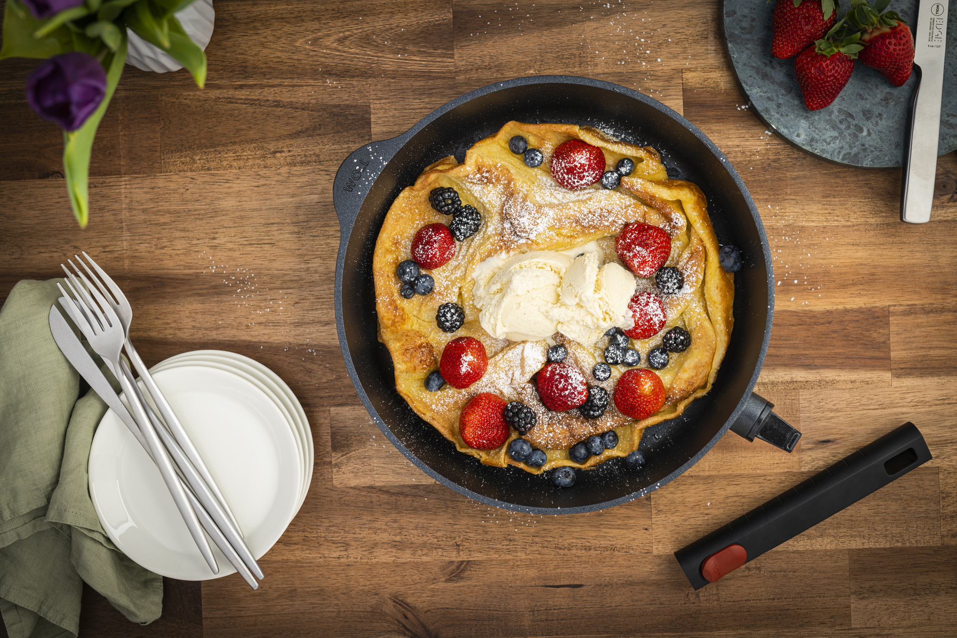 In einer runden Eco-Logic-QXR-Pfanne mit abgenommenem Stiel wurde ein Dutch-Baby-Ofenpfannkuchen gebacken. Auf diesem befinden sich frische Beeren, eine Kugel Eis und Puderzucker. Auf einem Brett oberhalb liegen frische Erdbeeren und ein Messer. Darunter