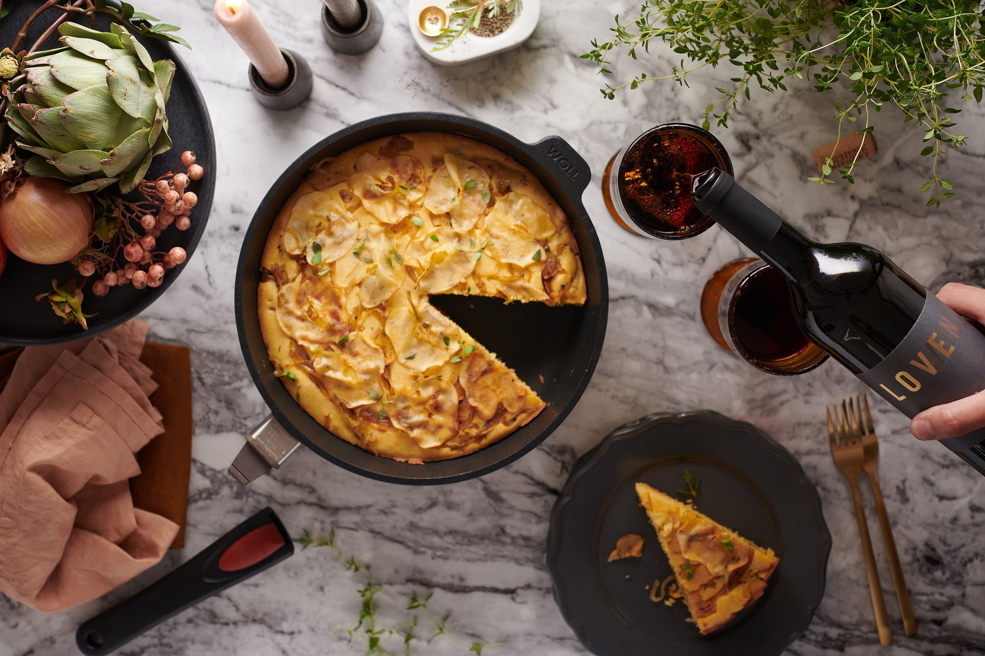Auf dem Tisch steht eine runde, heiße Titan-Best-Pfanne, in der sich ein frisch gebackenes Maisbrot mit karamellisierten Äpfeln und Zwiebeln befindet. Ein Stück davon wurde bereits auf einem Teller platziert. Drumherum stehen Weingläser, Zutaten und Kerze