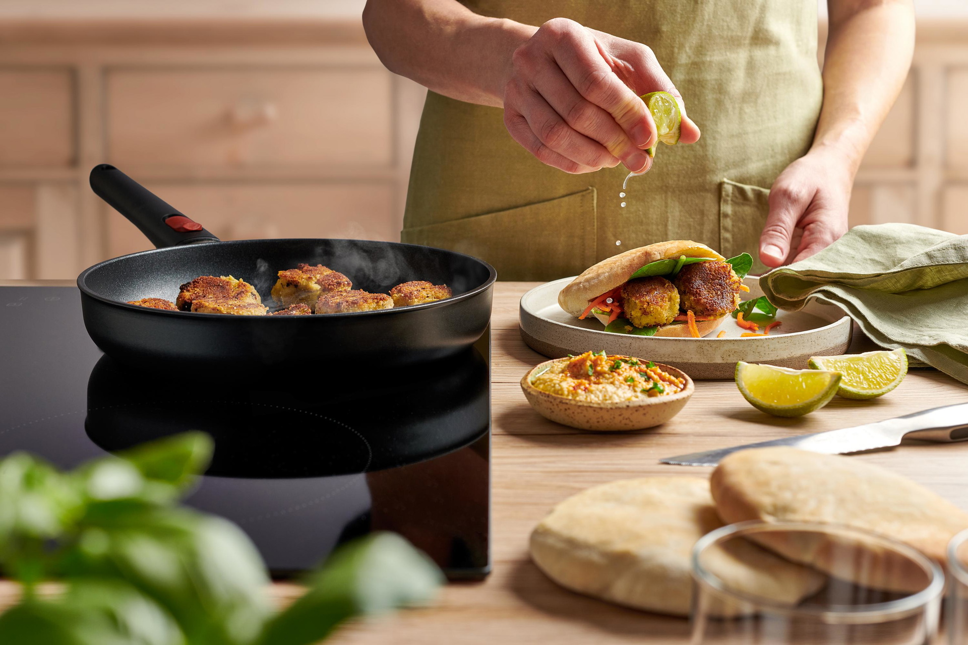 In einer runden Eco-Lite-QXR-Pfanne werden Falafel angebraten, während auf einem Teller ein gefülltes Fladenbrot liegt.