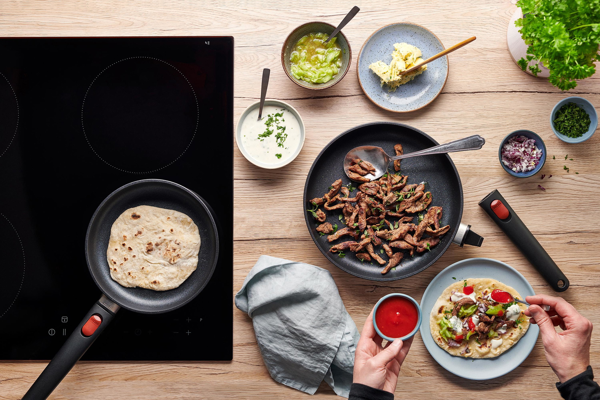 Eco Lite QXR Flachpfanne In der runden Eco-Lite-QXR-Pfanne werden Fladenbrote gebacken. Daneben liegt in einer Pfanne angebratenes Lamm und auf einem Teller ist Döner zubereitet.