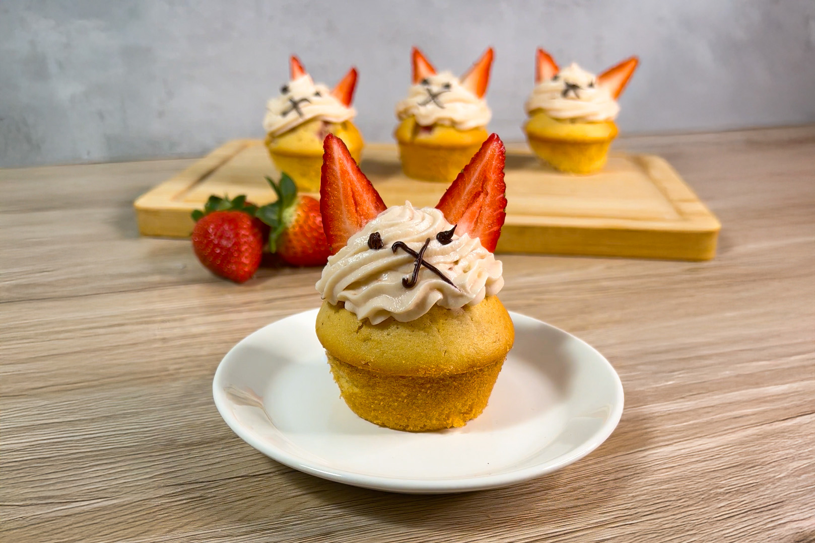 Auf einem Teller steht ein Cupcake mit Erdbeer-Frosting, dahinter sind auf einem Schneidebrett drei weitere zu sehen. Alle sind mit Zuckerstift und geviertelten Erdbeeren dekoriert.
