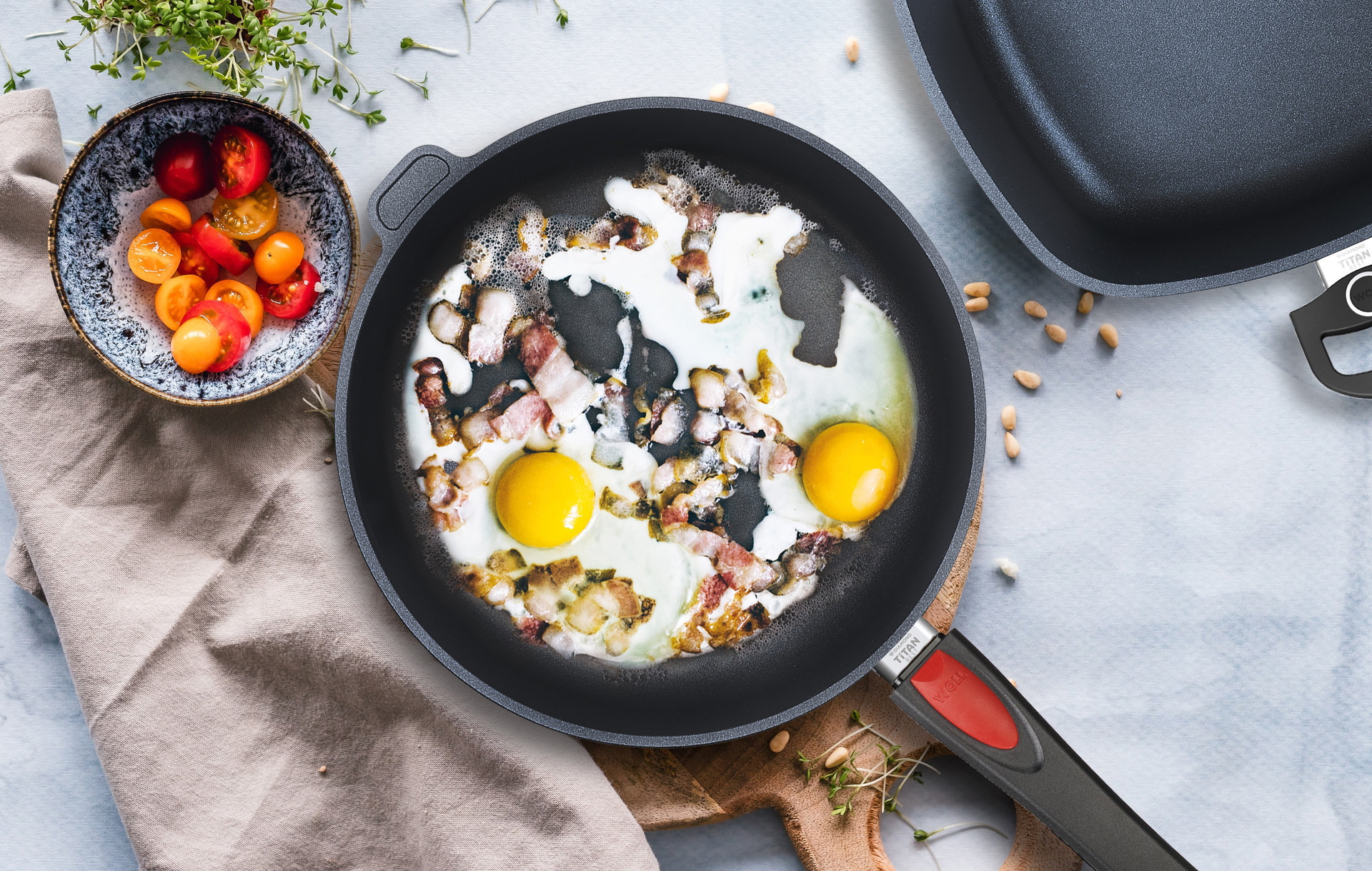 In einer runden, heißen Titan Best Pfanne mit abnehmbarem Stiel befindet sich frisch zubereitetes Spiegelei mit Speck, daneben steht eine Schüssel Tomatensalat.