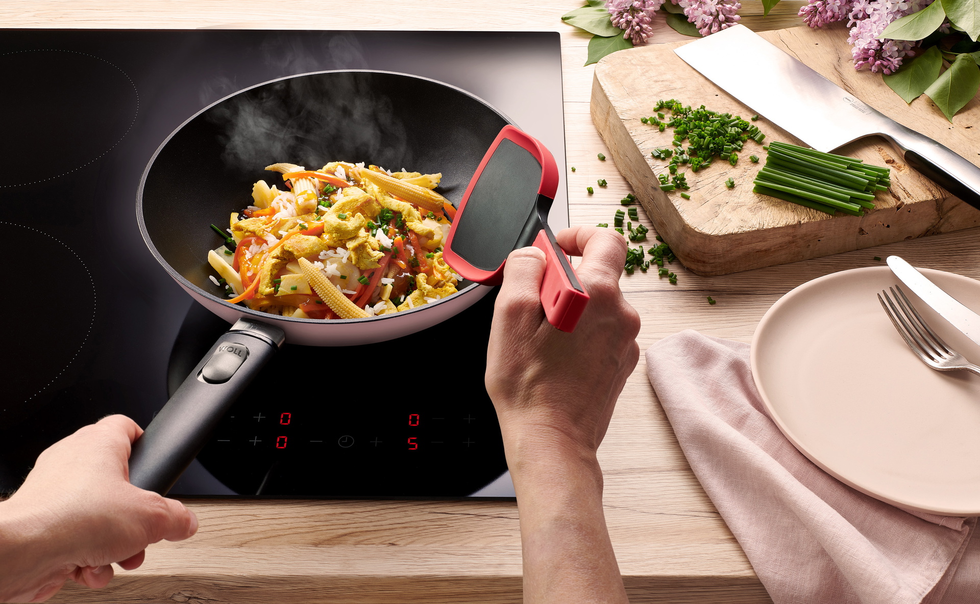 Eine runde, hellrosa Ecolour Wokpfanne steht auf dem Herd, mit einem Cock it wird ein dampfendes Gericht gewendet. Das Edge Chinesisches Kochmesser liegt daneben.